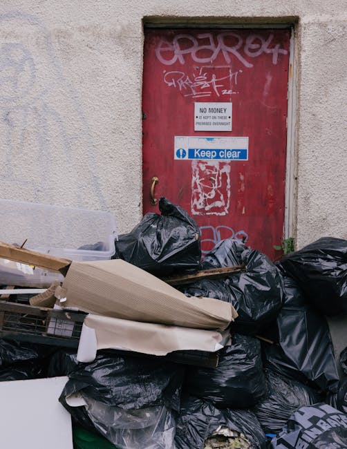 A red metal door set into a textured, off-white concrete wall, featuring graffiti tags and multiple stickers, including a 'Keep Clear' sign and a small 'No Money' notice. In front of the door, there is a large pile of black rubbish bags, some partially torn or bulging, with additional discarded materials such as flattened cardboard boxes, protective foam packaging, and pieces of cardboard, all stacked haphazardly on a concrete pavement, indicating a collection of waste or obsolete items awaiting removal. The scene suggests an area where unofficial or private waste disposal takes place, possibly as part of onsite rubbish clearance services by Rubbish Clearance Earls Court, highlighting the common need for professional rubbish removal solutions in urban environments.