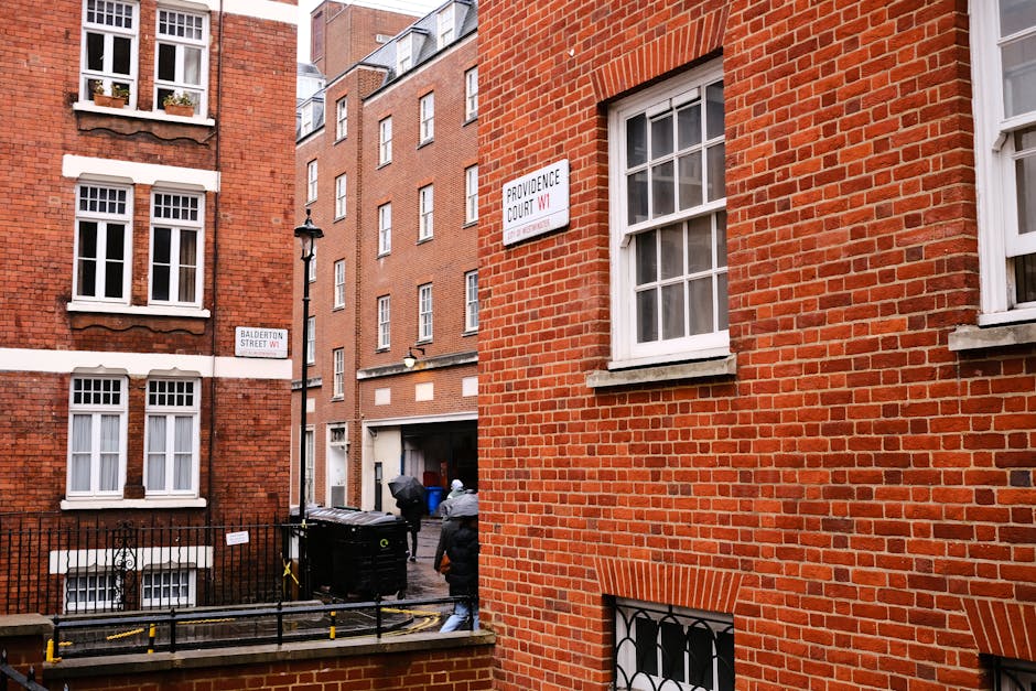 A narrow urban alleyway in a densely built residential area featuring three red-brick buildings with white framed sash windows. The building on the right has two visible windows on the upper floors, with white window sills and grid-patterned panes. A street sign labeled 'PROVIDENCE COURT SW1' is mounted on the wall of this building near the middle, and beneath it, another sign reads 'BALDERTON STREET SW1.' In the background, a modern brick building with multiple uniform windows partially obscures the view. At street level, a black wheelie bin is positioned next to a metal railing, and two individuals are loading or unloading rubbish bags nearby, suggesting ongoing rubbish clearance or private waste handling often associated with independent waste collection services. The scene is illuminated by natural daylight, with subtle shadows cast by the buildings and objects, creating a neutral and professional atmosphere focused on urban waste management activities.