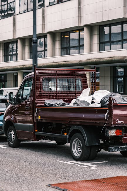 A small burgundy flatbed truck parked on an urban street in front of a modern building with large glass windows and a white concrete exterior. The truck's cargo area is filled with various black, grey, and white plastic bags containing waste or discarded materials, some of which appear to be tied or sealed. The truck features a metal mesh safety barrier behind the cab and has visible side mirrors and rear lights. The surrounding environment includes a paved road with street markings and a partially visible pedestrian crossing at the bottom of the image. Nearby, there is a lamppost and other parked vehicles, suggesting a typical city street scene. The image, associated with Rubbish Clearance Earls Court, depicts on-site waste collection or private rubbish removal, aligning with alternative waste handling services outside local authority disposal.