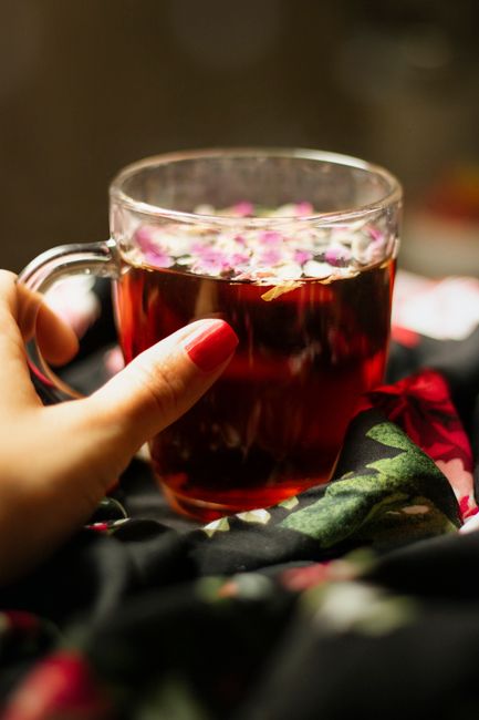 A close-up view of a transparent glass mug filled with dark amber or reddish herbal tea, topped with floating pink and white flower petals. The mug is held by a person's hand with neatly manicured nails painted in a soft pink shade. The background is softly blurred, with hints of dark fabric that features a floral pattern in shades of red, green, and black, possibly a decorative cloth or garment. The surface beneath the mug appears to be dark, contributing to a warm and cozy atmosphere. This scene exemplifies a moment of relaxation involving a beverage, which is relevant to private or alternative waste handling of domestic rubbish, such as disposing of used tea leaves and flowers, typical in settings managed independently without external collection services. The image conveys a calm, intimate setting associated with casual indoor rubbish management involving tea residues and floral garnishes, aligning subtly with principles of personal rubbish clearance and disposal.