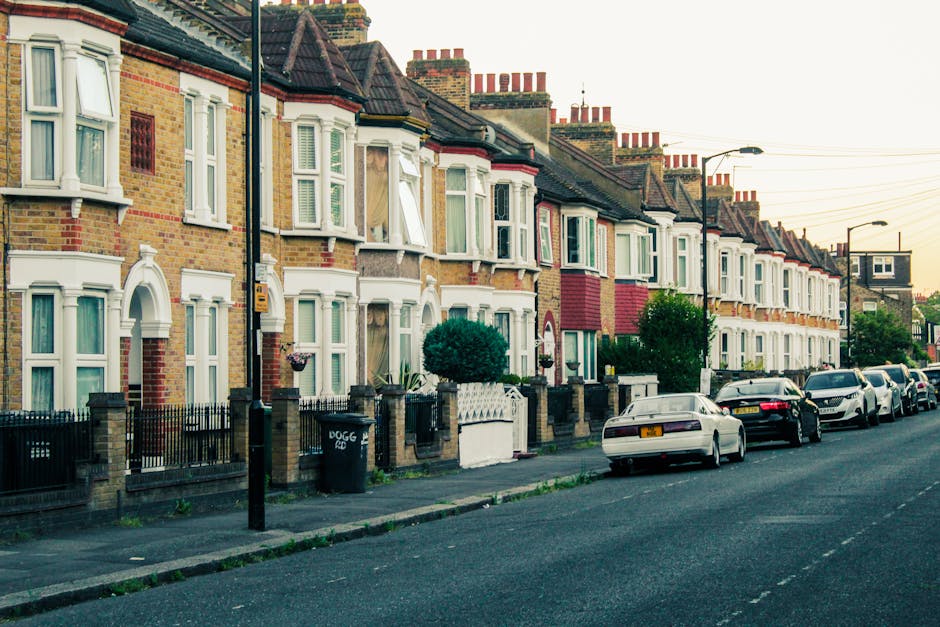 A row of traditional terraced houses with brick exteriors and ornate white window frames situated along a residential street in Earls Court. The houses have pitched roofs with multiple chimneys and are separated by small front gardens enclosed by low brick walls and wrought iron railings. Several houses feature bay windows, some of which are open, revealing curtains inside. The sidewalk in front is lined with street lamps, a utility pole, and a few small bushes or trees in front gardens. Parked along the curb are various vehicles, including a white car in the foreground and darker cars further back, all aligned parallel to the pavement. The scene is captured during late afternoon or early evening, with soft, natural light illuminating the buildings and the street. The environment suggests a quiet residential area that may benefit from private rubbish clearance services, exemplifying typical urban waste management needs for property upkeep and waste removal in the SW5 postcode area, managed by Rubbish Clearance Earls Court.