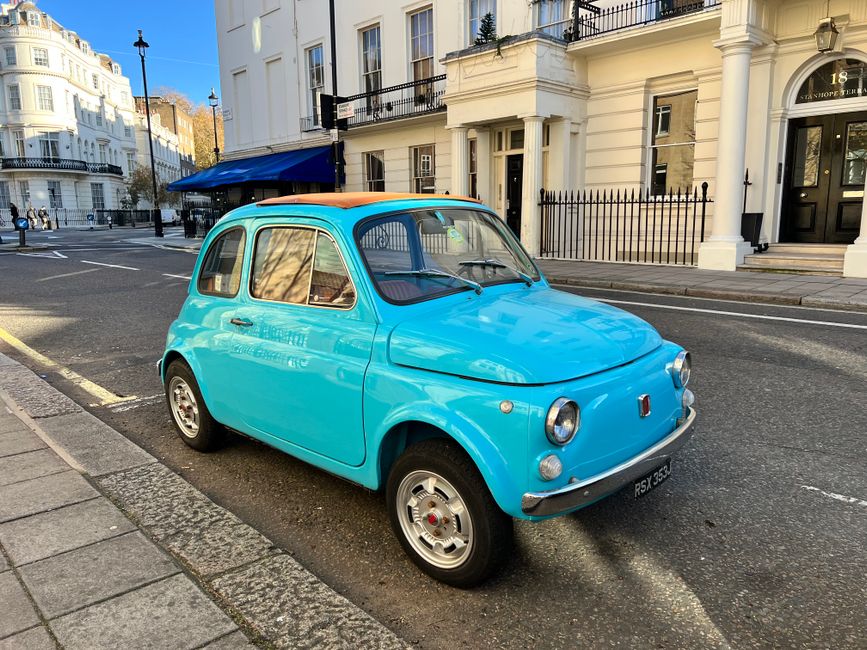 A vintage light blue Fiat 500 with a rounded compact body, small circular headlights, and a slightly elevated orange roof parked on a paved city street. The car's exterior has a shiny finish, with chrome accents around the headlights and on the bumpers. It is positioned near a curb with a concrete sidewalk, and behind it is a row of white, multi-story Victorian-style buildings featuring large windows, decorative moldings, and black wrought iron balconies. To the left, a modern black streetlamp lines the road, and in the background, additional vehicles and pedestrians can be seen walking along the pavement. The scene is illuminated by natural daylight, casting soft shadows on the car and surrounding environment. This setting illustrates a typical urban street scene, relevant to private vehicle storage and possible alternative disposal methods in the context of rubbish clearance and vehicle recycling, as often managed by companies like Rubbish Clearance Earls Court. The overall composition conveys a neat, well-maintained streetscape with a focus on classic vehicle preservation amidst city life.
