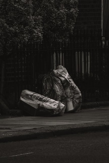 Two large black plastic rubbish bags, placed on the pavement near the edge of a street, appear to be filled with waste materials and are tied at the top. The bags are positioned close to a wooden fence and a tall, leafy tree, which cast shadows in the dim lighting. The pavement surface is smooth, and the street in the foreground is dark with a visible curb. The background shows a dark, possibly residential area with some faintly visible structure behind the fence, suggesting an environment where private waste disposal or on-site rubbish collection might be considered as alternative options to council rubbish rules, highlighting the importance of proper rubbish management services such as those offered by Rubbish Clearance Earls Court.