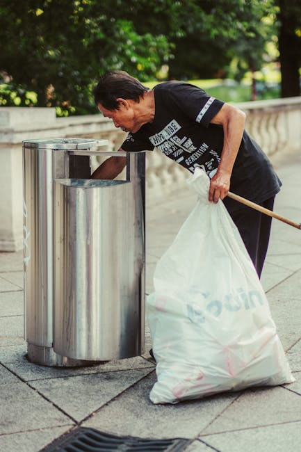 A middle-aged man with dark hair dressed in a black T-shirt with white and blue text, is seen bending over to place a plastic bag filled with rubbish into a tall, stainless steel litter bin at an outdoor location lined with paving stones. The bin is cylindrical with a shiny, reflective surface and a rectangular opening at the top. The man holds a wooden stick or litter picker in his left hand, which is resting on his hip, while his right hand is guiding the bag into the bin. Behind him, a low, carved stone or concrete wall separates the walkway from a lush area with dense green foliage, including trees and bushes. The scene appears to be part of a routine waste collection or on-site rubbish removal, typical of independent waste handling services like those offered by Rubbish Clearance Earls Court, supporting private rubbish disposal of commercial or residential waste in the area. The natural daylight brightly illuminates the scene, emphasizing the metallic texture of the bin and the casual, everyday activity of rubbish management in an urban setting.