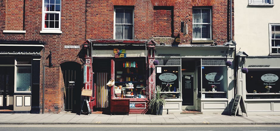 A row of small retail units and cafes along a city street, with a focus on a shop displaying various items including colorful signs, jars, and decorative objects beneath an awning. The shop's open facade reveals a mixture of retail displays and shelves filled with products. To the right, there are large front windows with flower pots on the windowsills, and a narrow door in the center. The adjacent buildings are constructed with red brick and painted brick materials, with visible windows and architectural details. The pavement in front of the shops is clean and free of rubbish, with a clear roadway beyond. The overall scene portrays an urban retail environment, reflecting a setting where independent waste collection or on-site clearance might be relevant for local businesses or property owners seeking alternative rubbish removal options, as handled by companies like Rubbish Clearance Earls Court.